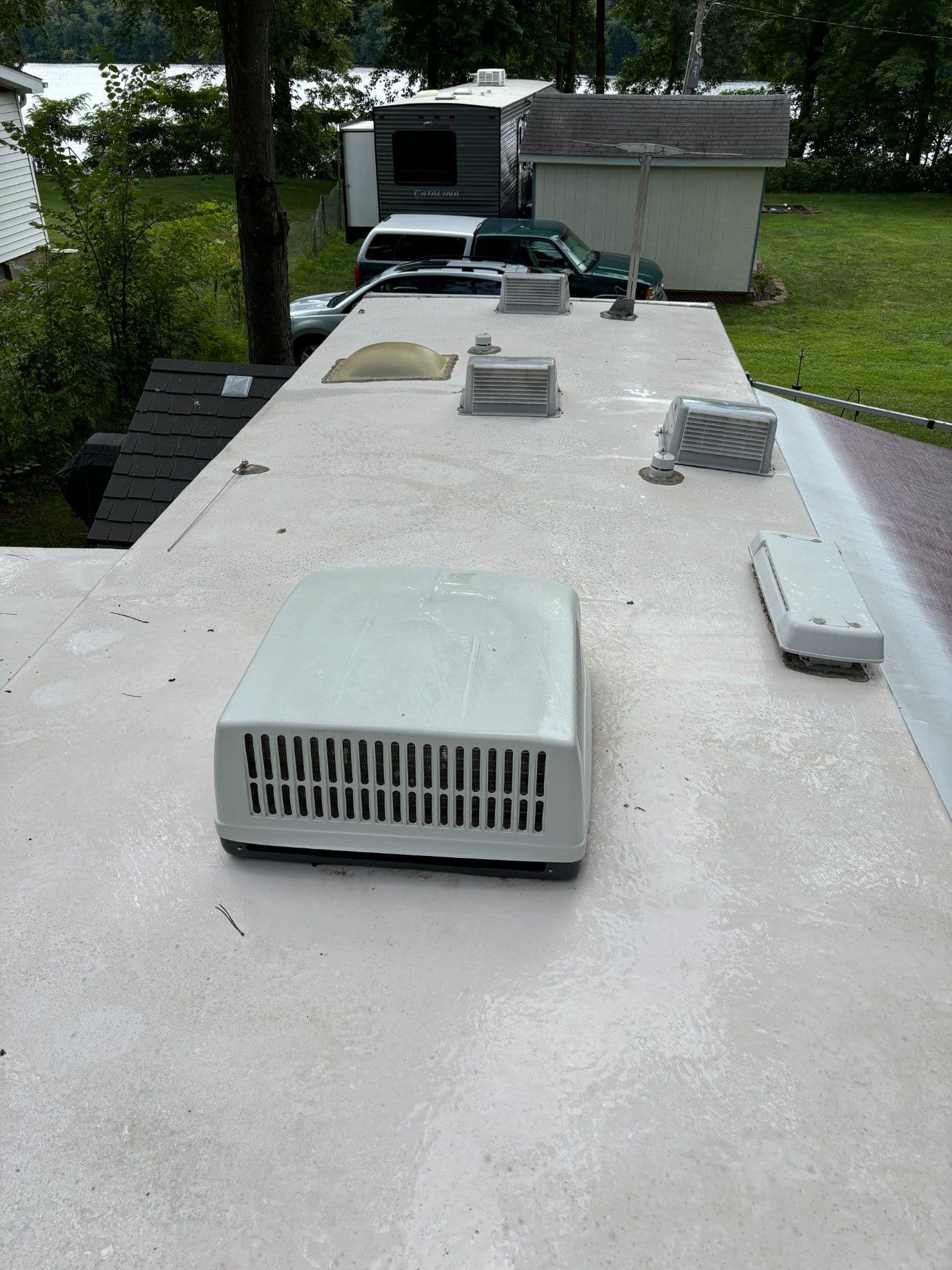 Roof of an RV with vents, air conditioner, and surrounding trees and vehicles.