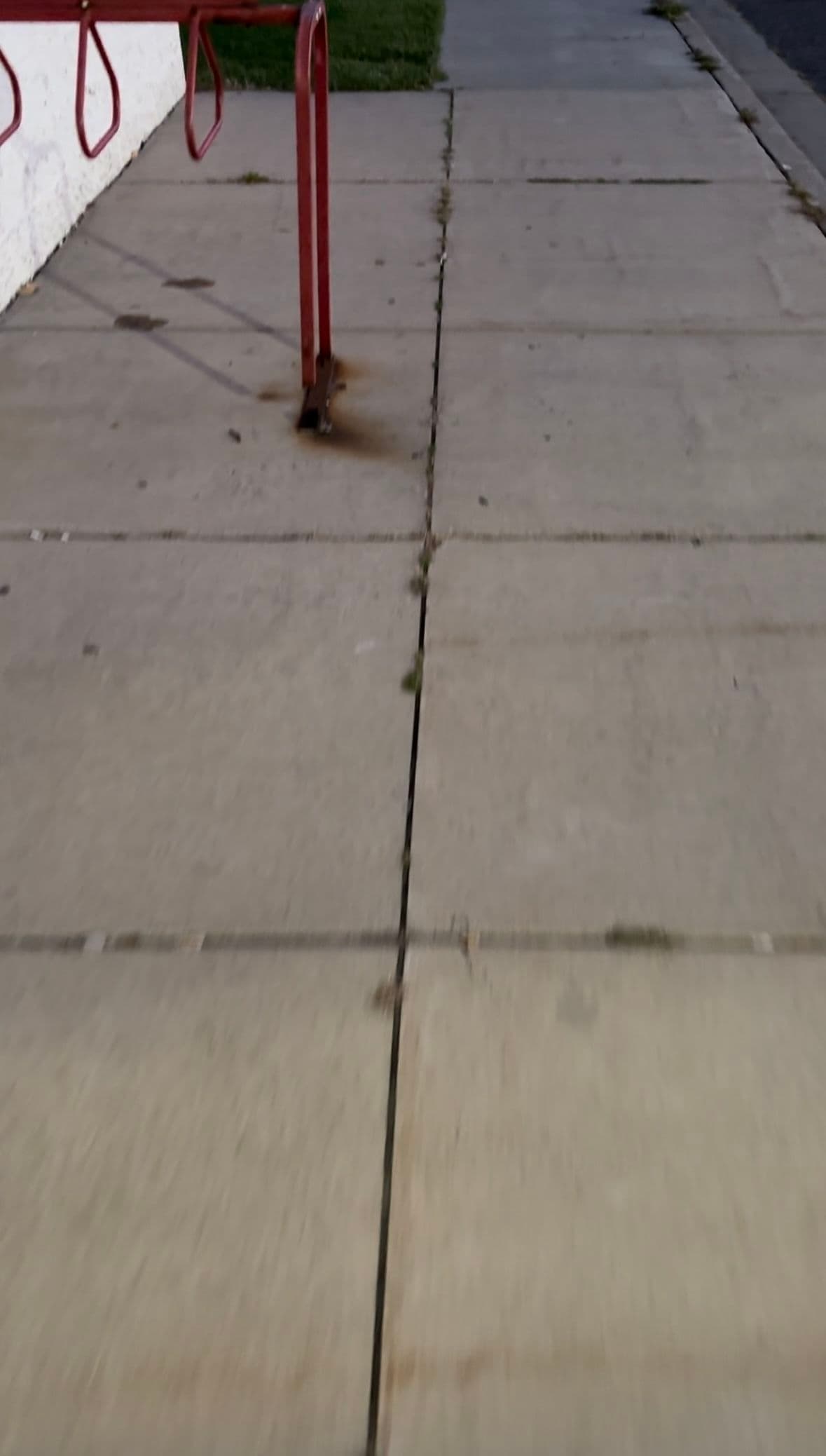 Sidewalk with visible cracks and a red bike rack against a white wall.