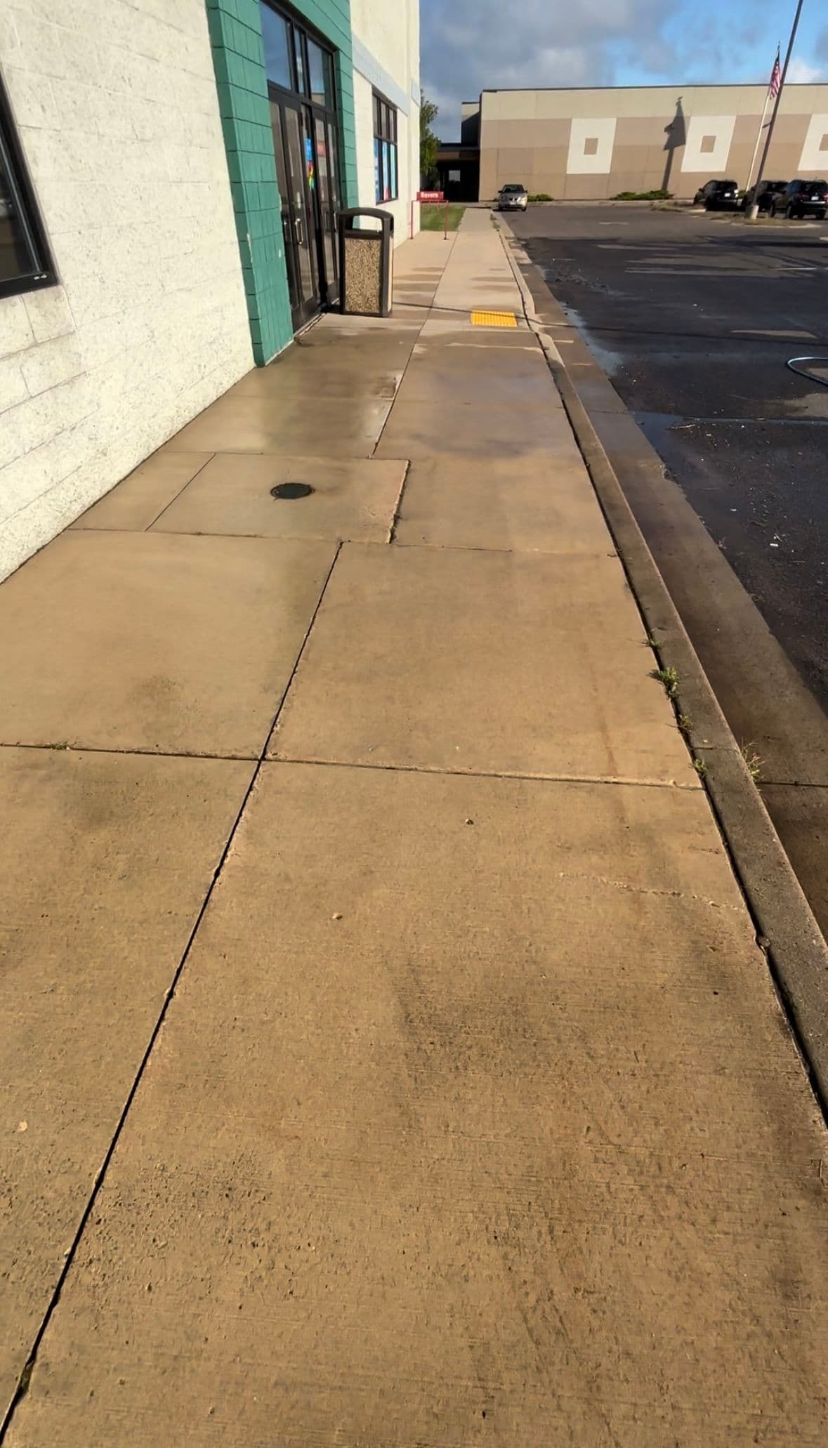 Wet sidewalk leading to a building entrance with trash can, clear blue sky in background.